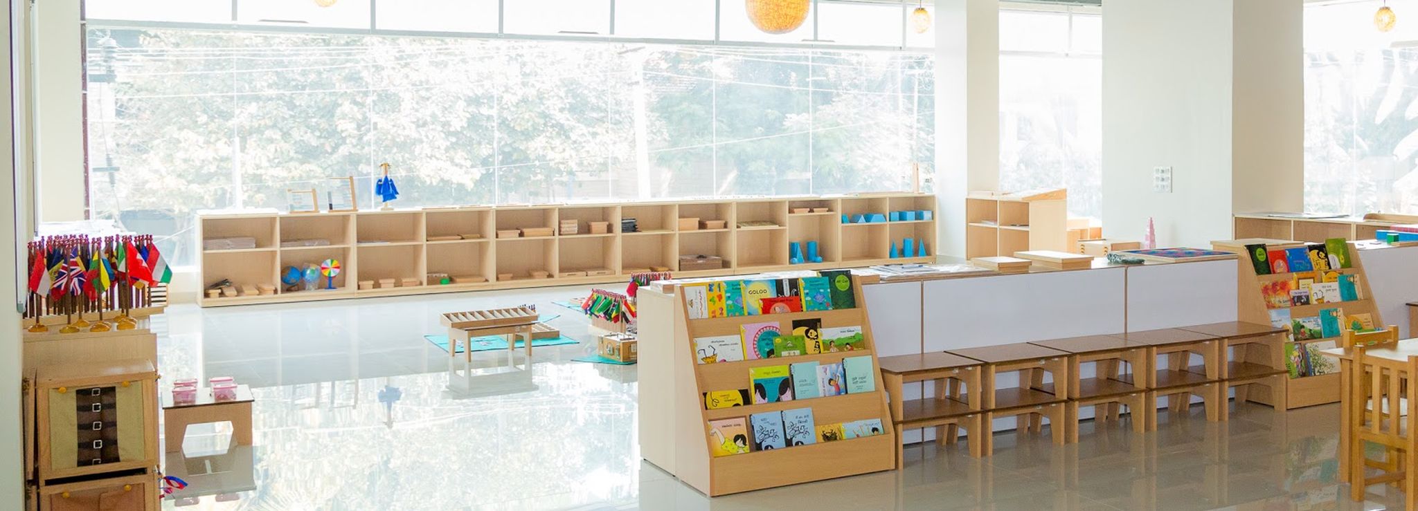 An empty Children&rsquo;s House in daylight &mdash; low shelves along a window wall, book displays at child height, and small tables and chairs waiting for the children.