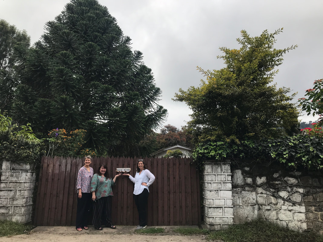 The wooden gate of Rose Bank in Kodaikanal, its painted name plate set between whitewashed stone pillars under tall trees. Three visitors stand at the gate.